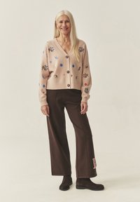 Hele roosa kootud kardigan, millel on värvikas lilleline ja ringikujuline tikand, V-kaelusega, esiküljel nööbid ja soonikkoes varrukasuu. Sobitatud pruunide laiade pükstega.