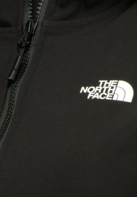 Giacca nera con zip realizzata in tessuto liscio, caratterizzata da un logo bianco di "The North Face" sul lato sinistro superiore. Il cursore della zip è nero.