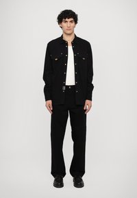 Camisa negra con dos bolsillos en el pecho y botones dorados, combinada con pantalones negros y zapatos negros. Camiseta blanca lisa visible.