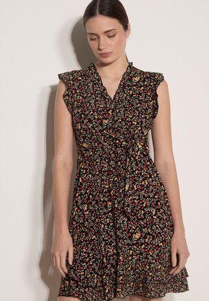 Kobieta ubrana w czarną sukienkę bez rękawów z drobnym kwiatowym nadrukiem w kolorach czerwonym, żółtym i zielonym, z falbankami na ramionach i dekoltem w kształcie litery V.