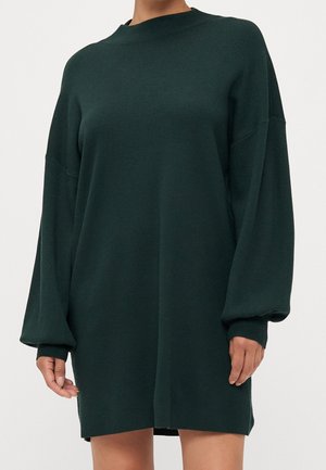 Femme portant une robe pull en maille vert foncé à manches longues, avec un col montant et des poignets côtelés.
