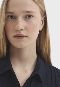 Jeune femme aux longs cheveux blonds et aux yeux gris portant une chemise sombre à col, regardant droit devant elle sur un fond clair.