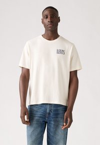 Balts kokvilnas t-kreklis ar apaļu kakla izgriezumu, īsām piedurknēm un zilu "LEVI'S" logo kreisajā krūtīs, saskaņots ar ziliem džinsiem.