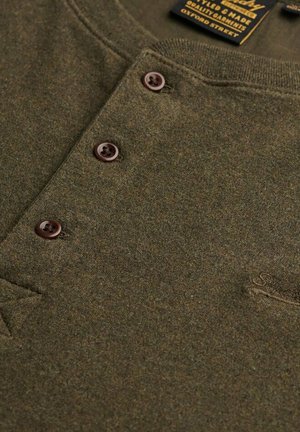 Oliven grøn henley skjorte med tre brune knapper og en synlig etiket nær halsudskæringen, der viser mærke- og kvalitetsoplysninger.