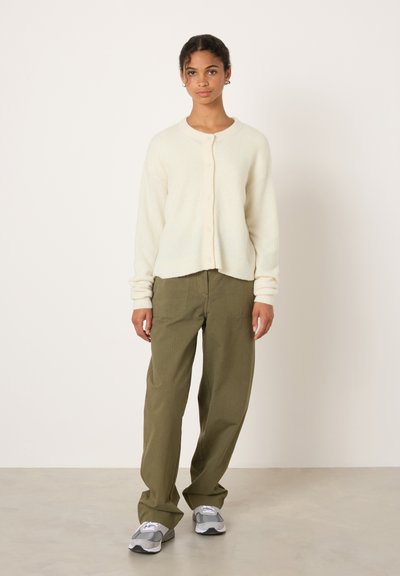 Cardigan crème avec fermeture à boutons, coupe décontractée et poignets côtelés. Associé à un pantalon large vert olive et des baskets grises.
