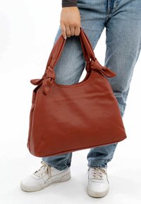 Bolsa tote de cuero marrón con acentos de asas anudadas, textura suave y un diseño espacioso, sostenida por una persona con jeans desgastados y zapatillas.