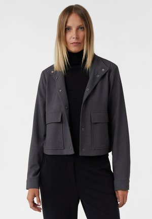 Frau mit glattem blonden Haar, die eine dunkelgraue, geknöpfte Jacke über einem schwarzen Rollkragenpullover und schwarzen Hosen trägt, vor einem einfarbigen hellen Hintergrund.
