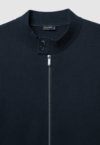 Maglione in maglia blu scuro con colletto alto, due bottoni neri vicino al collo e una cerniera argentata che scorre verticalmente lungo il centro.
