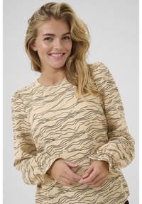 Ljusbeige blus med ett vågigt svart mönster. Har långa, sammanknölade ärmar och rund halsringning. Mjukt, texturerat tyg.