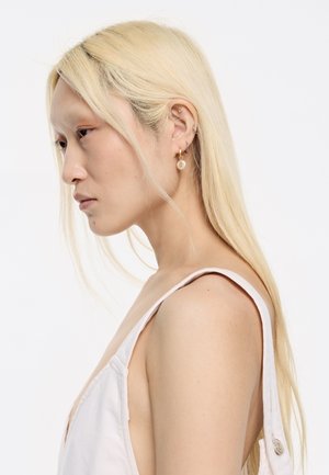 Blonde Frau mit langen Haaren in einem hellrosa ärmellosen Oberteil, die mehrere goldene und silberne Ohrringe trägt und nach unten blickt, vor einem weißen Hintergrund.