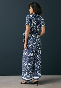 Blommig byxdress i marinblå med vita blommor, korta ärmar, vid ben-design och knytning vid midjan. Texturerat tyg och randig fåll.