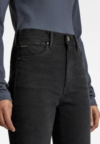 Persona vestida con jeans de cintura alta en gris oscuro y una blusa ajustada de manga larga y tejido acanalado en gris, con las manos relajadas a los lados.