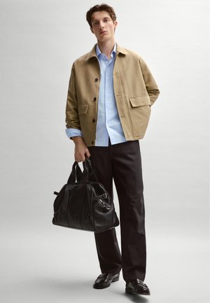 Muž s béžovou bundou, světle modrou košilí, černými kalhotami a černými botami drží černou koženou sportovní tašku na bílém pozadí.