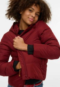 Mørkerød dunjakke med hette, glidelås foran, ribbede mansjetter og to sidelommer, brukt over en svart topp og blå jeans. Krøllete hår.