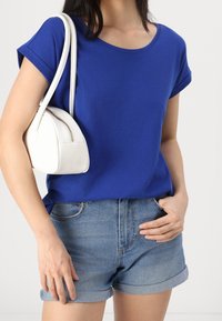 Nainen, jolla on sininen lyhythihainen paita ja kääritty farkkushortsit, kantaa valkoista pientä käsilaukkua olallaan yksiväristä taustaa vasten.