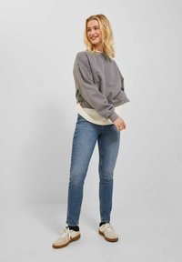 Grauer Sweatshirt mit einem cropped Design über einem cremefarbenen Langarmshirt, kombiniert mit engen blauen Jeans und beigen Sneakers.