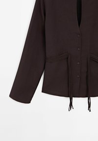 Chemise à manches longues marron foncé boutonnée avec encolure en V, ornée de détails en cordon décoratif et de pompons à la taille sur fond blanc.