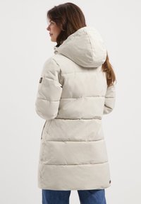 Vrouw in een lange, beige gewatteerde winterjas met capuchon, die zijwaarts staat met handen in de zakken tegen een effen achtergrond.