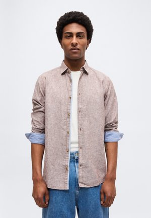 Homme aux manches retroussées portant une chemise marron clair à boutons sur un t-shirt blanc et un jean bleu, debout devant un fond clair uni.