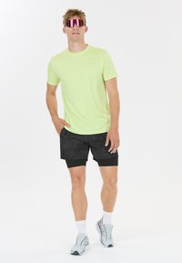 Ljusgrön kortärmad träningströja med svarta mönstrade shorts. Modellen bär reflexiva rosa solglasögon och grå löparskor.