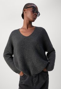 Grauer Strickpullover mit V-Ausschnitt,落肩 und langen Ärmeln. Weiche Textur; kombiniert mit einem schwarzen Rock. Modell trägt überdimensionale Sonnenbrille.