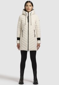 Elfenben isoleret jakke med chevron quiltning, frontlynlås, hætte med snøre, sorte manchetter og lommer. Modellen har sorte leggings og støvler på.