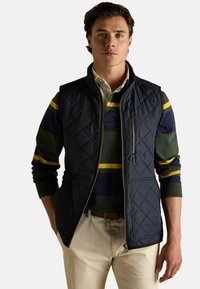 Námornícky prešívaný vest s zipsom, obohatený o šikmé vrecká. Nosený na pruhovanej dlhý rukáv zeleno-námorníckej a žltej košeli. Krémové nohavice.