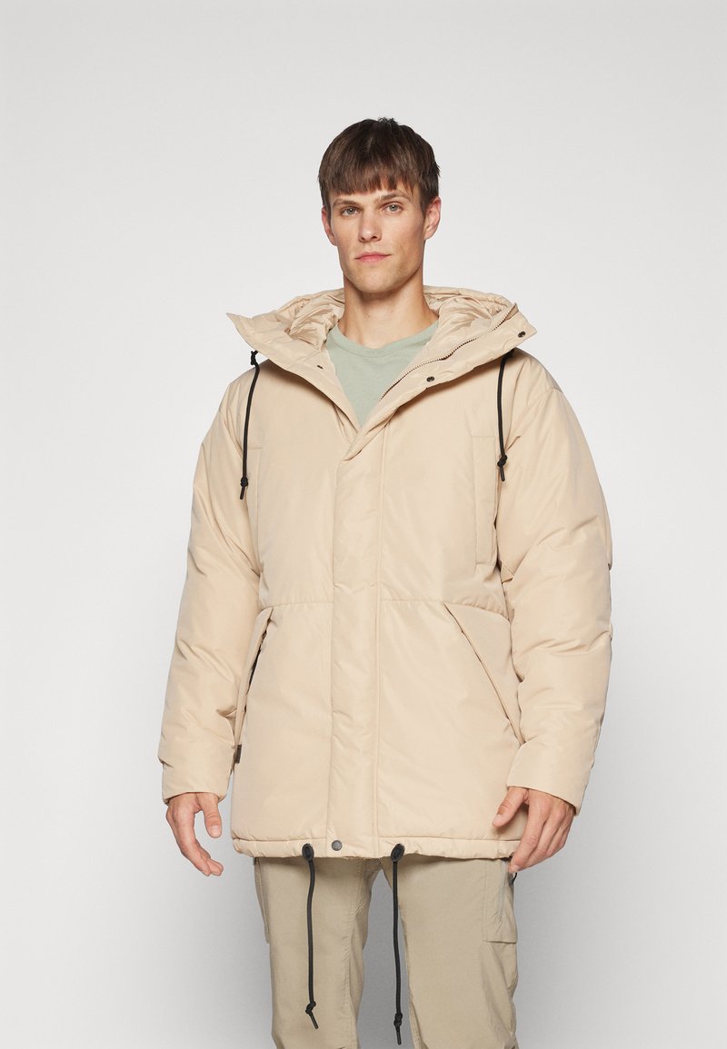 Beige boblejakke med hette, med glidelås, justerbare strammere og sidelommer. Glatt tekstur og polstret design.