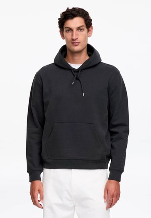 Mężczyzna ubrany w czarną bluzę z kapturem i kieszenią z przodu oraz białe spodnie, stojący przodem na białym tle.