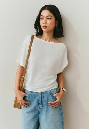 Jeune femme aux cheveux noirs porte un haut blanc épaules dénudées et un jean bleu, portant un sac bandoulière marron, debout contre un mur uni.