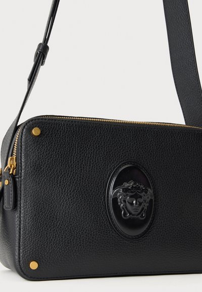Μαύρη υφασμάτινη δερμάτινη τσάντα crossbody με χρυστό φερμουάρ και τρουκ, με μαύρο ανάγλυφο έμβλημα κεφαλιού Μέδουσας στο μπροστινό μέρος.