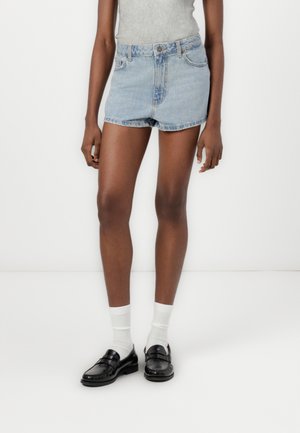 Lyseblå denimshorts med høj talje og frynset kant, parret med sorte loafers og hvide sokker, vist mod en neutral baggrund.