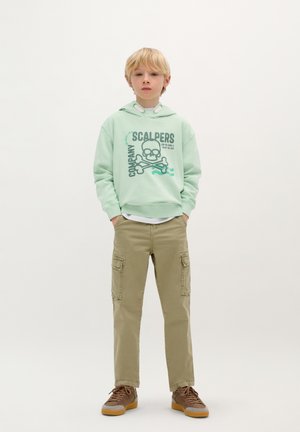 Mintgroene hoodie met een doodskop en botten grafiek, gecombineerd met lichtbeige cargobroek met zijzakken en bruine sneakers.