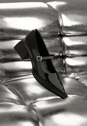 Black patent leather high-heeled shoe with a pointed toe, ankle strap, and chunky heel, set against a shiny metallic background.