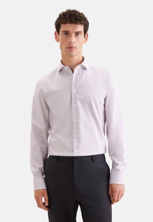 Uomo con camicia chiara a righe verticali e pantaloni scuri formali, in piedi davanti a uno sfondo uniforme.