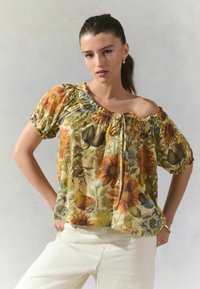 Blusa floral sin mangas con base amarillo-verde, decorada con girasoles y otras flores, mangas con puños elásticos y detalle de lazo en la parte frontal.