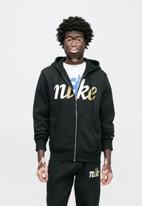 Čierna mikina na zips s kapucňou so zlatobielym logom „nike“. Kombinovaná s čiernymi teplákmi s rovnakým logom. Hladká textúra látky.