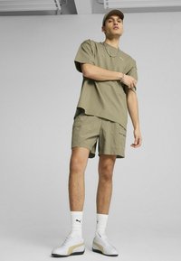 Maslinasto zelena prevelika majica kratkih rukava s odgovarajućim cargo šortsevima. Bijele tenisice s zlatnim detaljima i bijele čarape s logotipom.