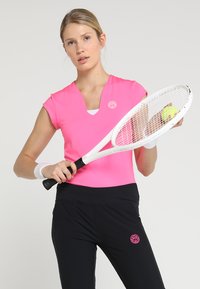 Top sportivo fitted di colore rosa fluo con maniche corte e scollatura a V, abbinato a pantaloni sportivi neri con un piccolo logo sulla vita. Il giocatore tiene una racchetta da tennis.