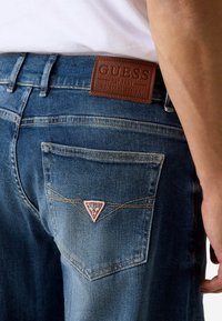Jeans di denim blu con una toppa in pelle marrone chiaro con la scritta "GUESS" in rilievo, un logo triangolare bianco e rosso sulla tasca posteriore e cuciture a contrasto.
