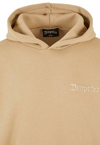 Beige hoodie with a front pocket and a drawstring hood. Features white embroidered logo on the left chest. Soft, smooth fabric texture.