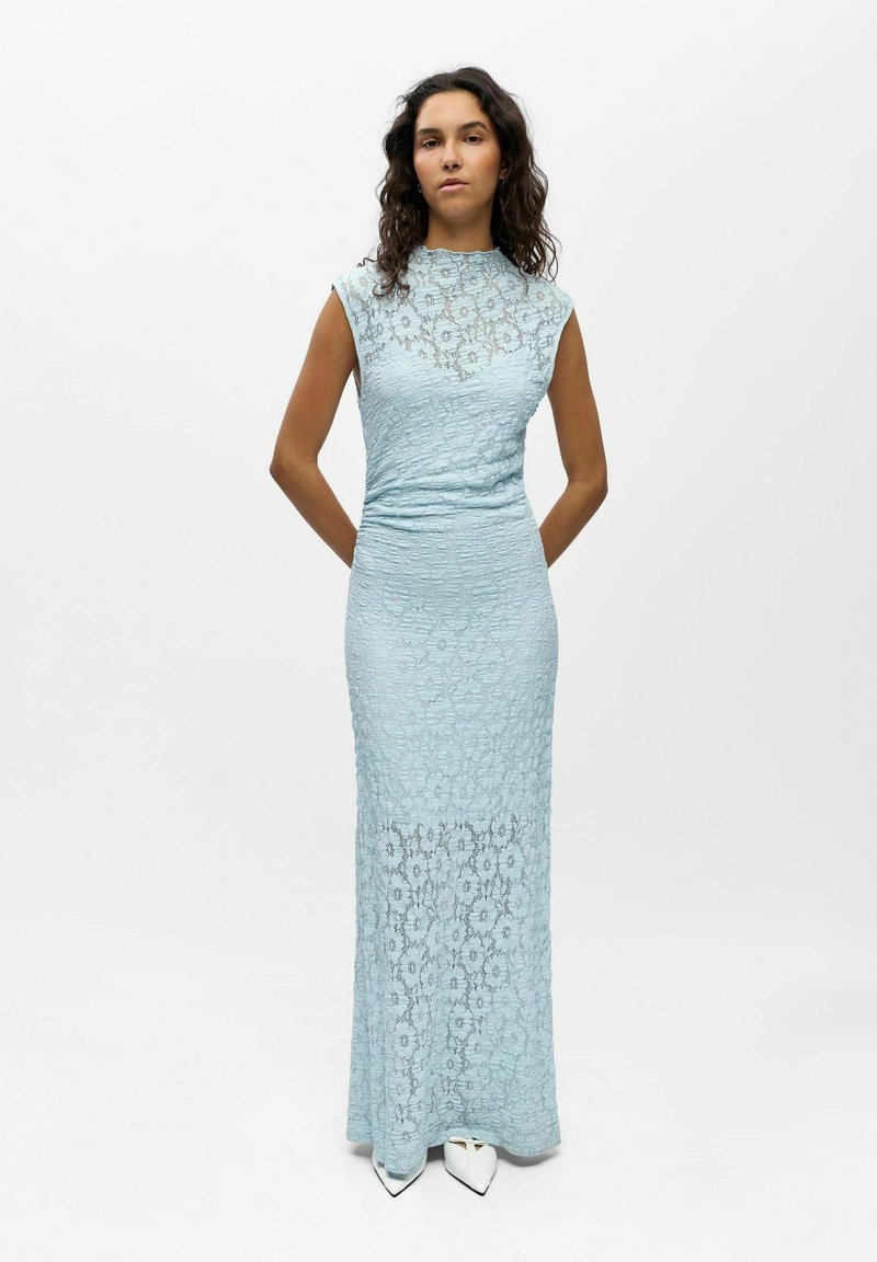 Femme aux cheveux bouclés portant une robe sans manches en dentelle florale bleu clair avec un col montant et des chaussures blanches à bouts pointus, debout devant un fond blanc.