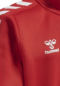 Giacca sportiva rossa in tessuto liscio con motivi a chevron bianchi sulle spalle e un logo con un design di ape sul petto.