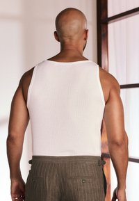 Débardeur blanc ajusté à texture côtelée, porté avec un pantalon marron à fines rayures, équipé de passants pour ceinture et de boutons décoratifs. Vue de dos.