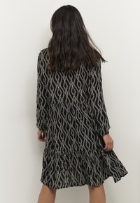 Robe noire à manches longues, présentant un motif abstrait blanc. Texture fluide, taille froncée, et longueur au genou.