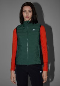 Femme aux cheveux raides et foncés porte une doudoune verte par-dessus une chemise rouge à manches longues et un pantalon noir avec des logos « 4F », debout devant un fond gris.
