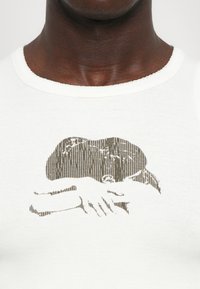 Λευκό αμάνικο μπλουζάκι από βαμβάκι, με μια υφασμάτινη καφέ γραφική απεικόνιση χεριών και καπέλου, διακοσμημένο με λεπτές κάθετες γραμμές.