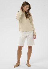 Haut beige à manches longues, short blanc et ballerines tissées beige. Le haut a une coupe ample ; le short arrive au genou avec un ourlet net.