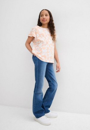 Top corto pesca con maniche corte e motivo floreale bianco, abbinato a jeans blu e sneakers bianche. Il modello è in posa rilassata.