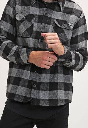 Mand iført sort og grå ternet flannelskjorte, der knapper manchet på venstre håndled.